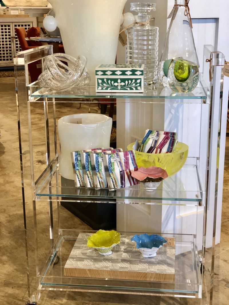 Clear acrylic and glass bar cart filled with various items, including cups, bowls, and vases.