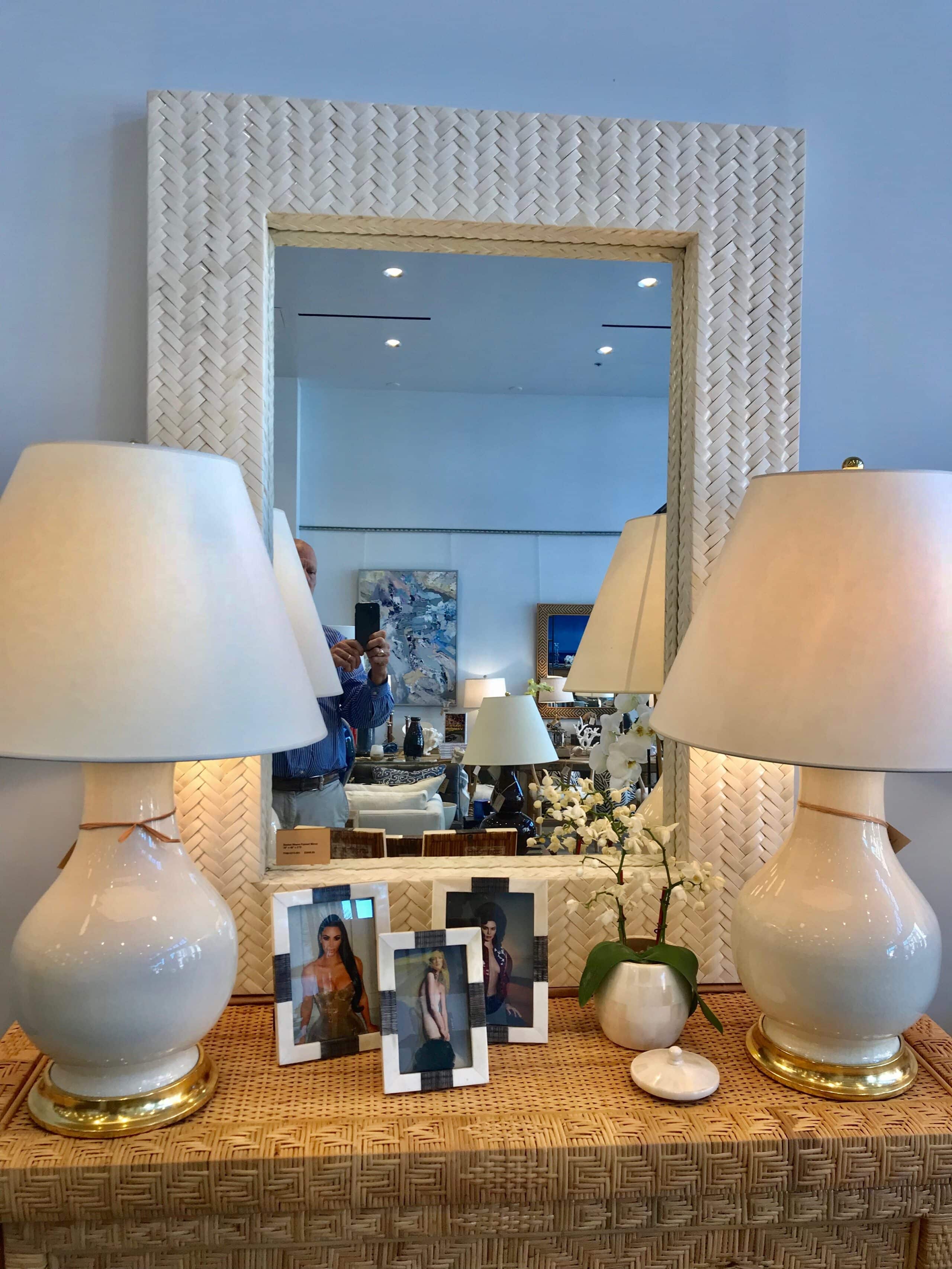 Basket weave mirror with bone frame, placed on a table with two lamps and a framed photo.