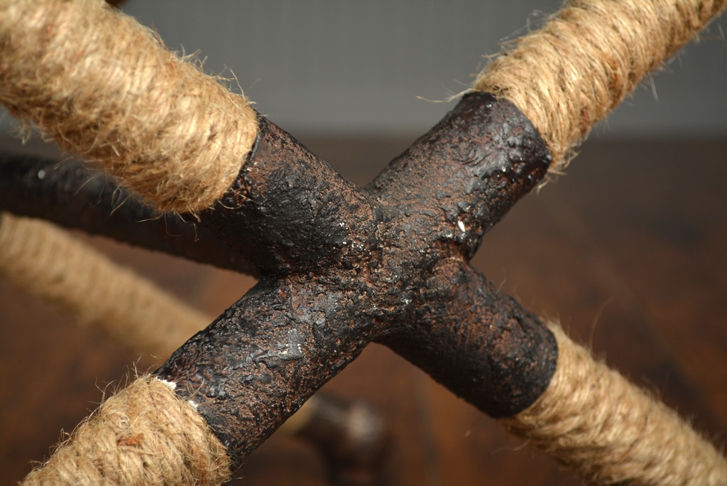 A rope stool with a cross design, featuring blue and white stripes on its seat and a rustic iron frame.