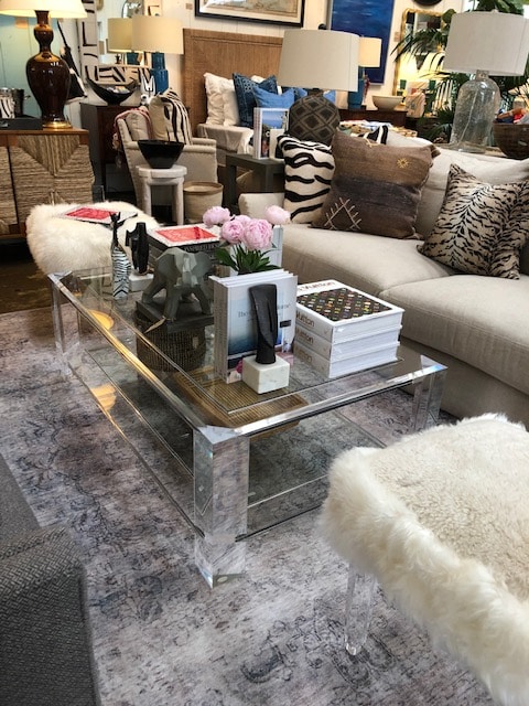 A clear glass coffee table with a bottom shelf, situated in a well-decorated living room.