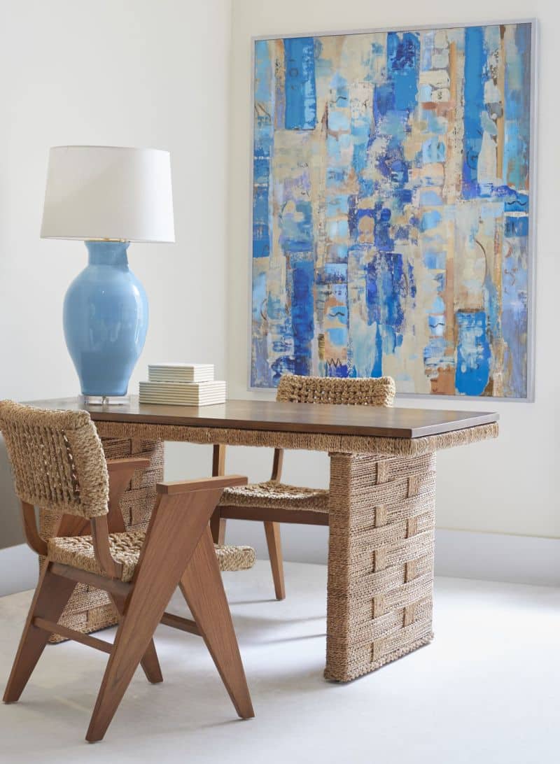 A blue vase on a table with a woven base and teak top, set against a colorful abstract painting.