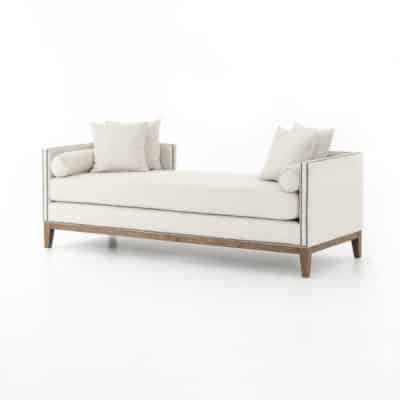 A cream upholstered daybed with wooden legs and brass nailheads, placed on a white surface.