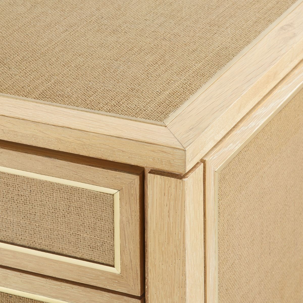 A wooden nightstand with three drawers, featuring a natural oak finish and jute panels on the sides, with brass handles.