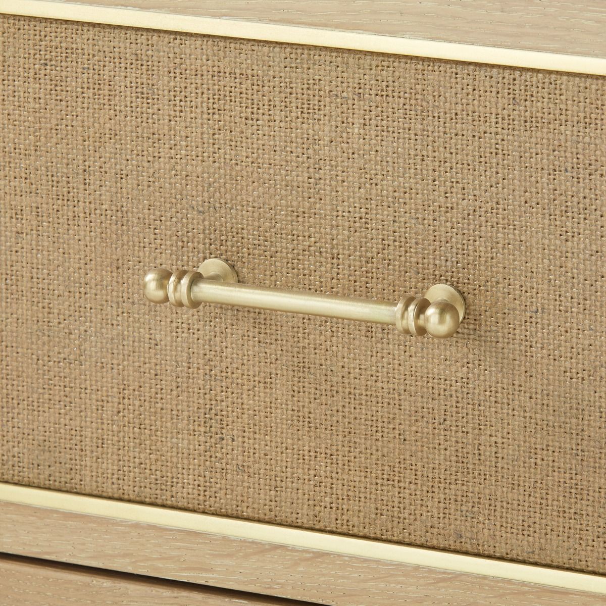 A three-drawer side table with a natural oak finish and woven jute panels, featuring brass handles and set against a neutral background.