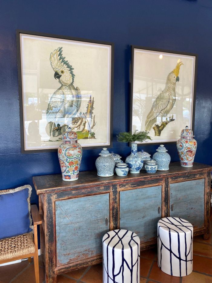 Two framed bird paintings on a wall above a console table a blue wall in the background decorative vases and round ottomans
