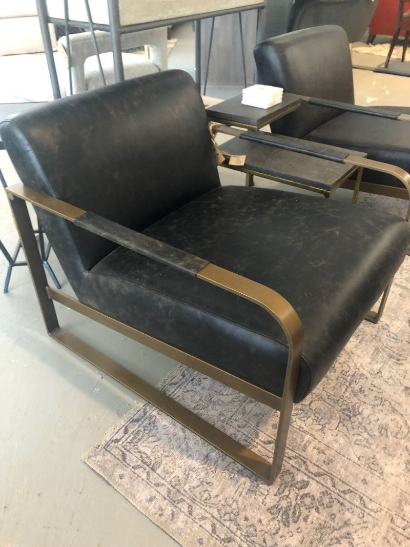 Ebony leather arm chair with brass frame, featuring tufted backrest and brass legs on a black carpeted floor.