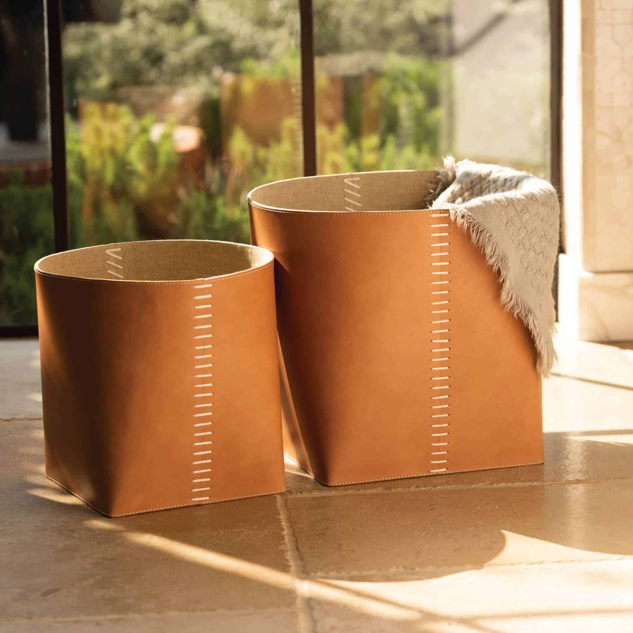 Set of two leather baskets with stitched detail, positioned next to each other on a tiled floor in front of a window.