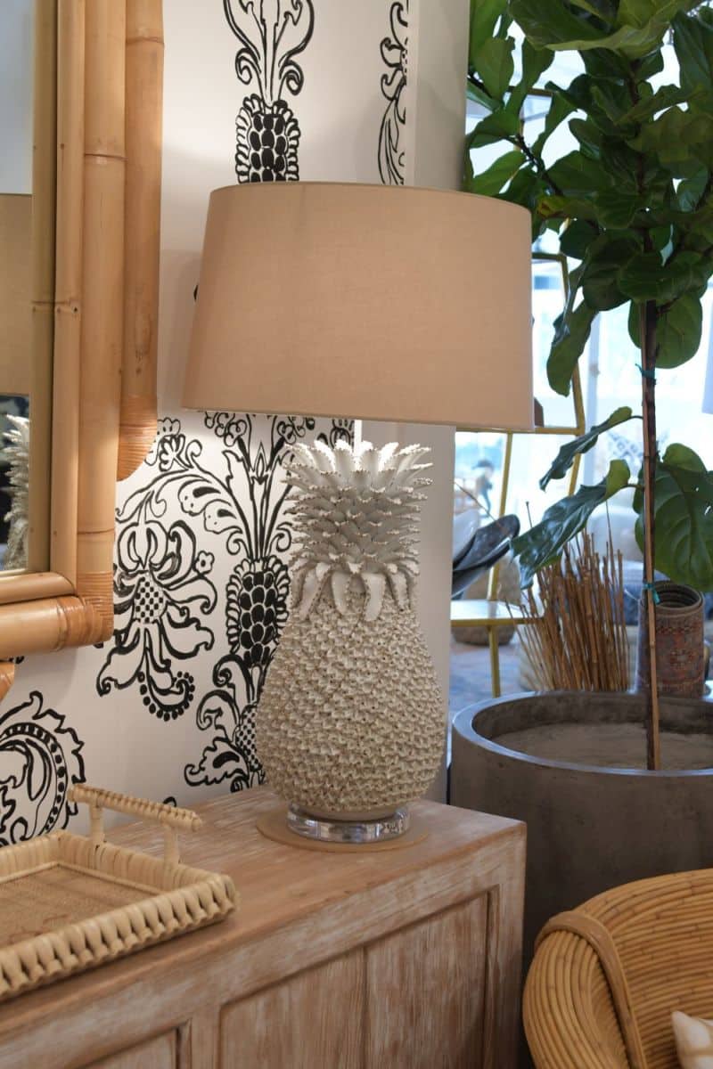 Large white pineapple lamp on table with bamboo lamp shade, side view, in room with potted plant and framed wall art.