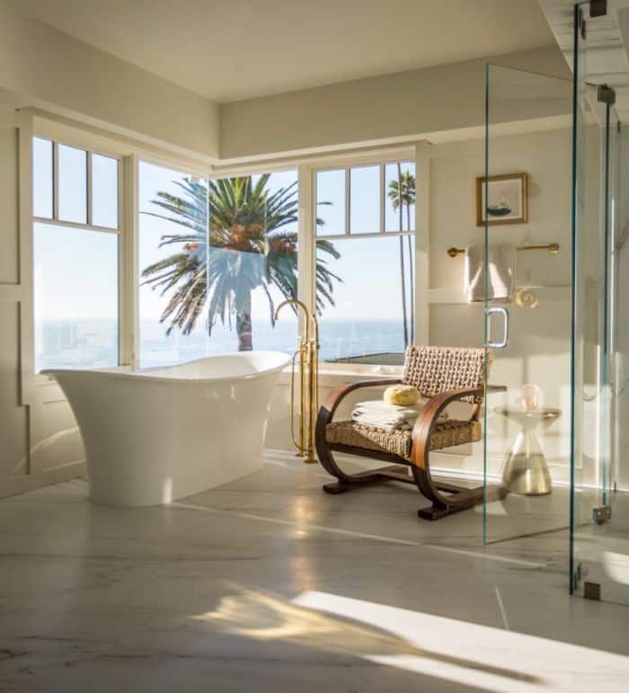 Luxurious modern bathroom with large window, white soaker tub, and sliding glass door leading to patio with palm tree.