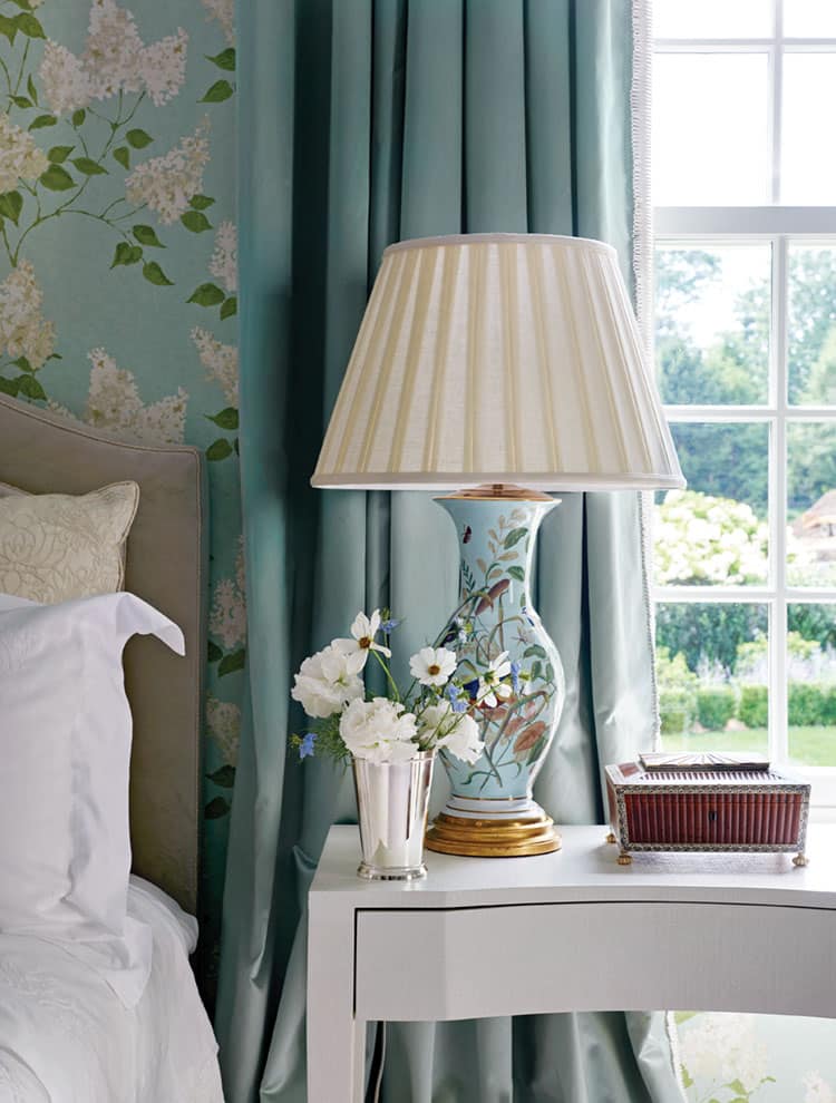 A bedside table with a blue and white striped curtain a lamp with a cream shade and a vase with flowers on