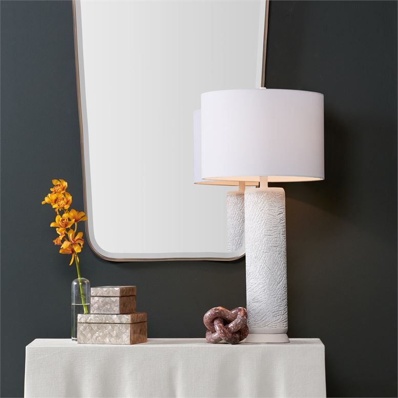 A tall modern lamp with a cylindrical base standing on a table against a dark wall background The lamp features a white