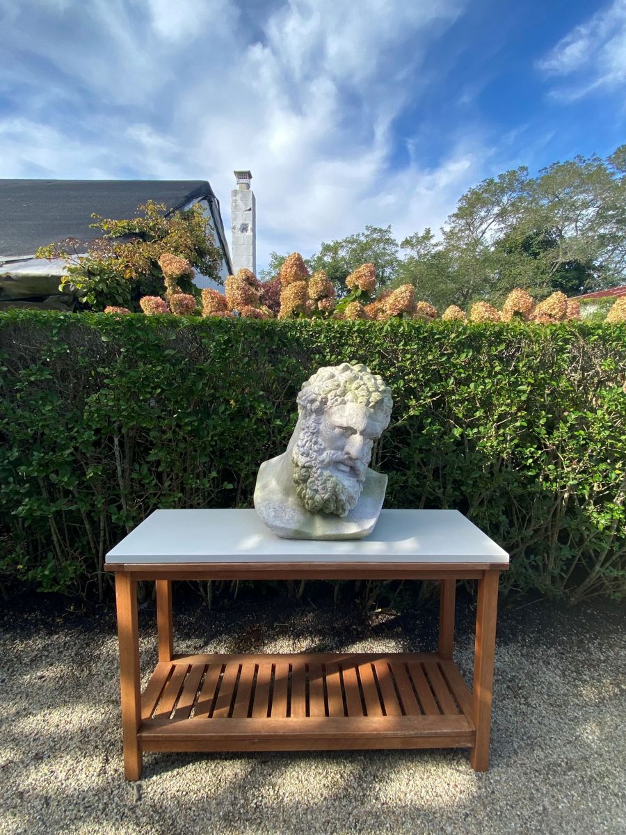 A white garden statue depicting Heracles set on a wooden bench against a backdrop of bushes and hedges showcasing its intricate details