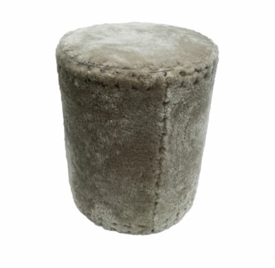 A close-up image of a cylindrical ottoman with a soft shearling upholstery and a pewter nailhead trim available in dusty beige ivory
