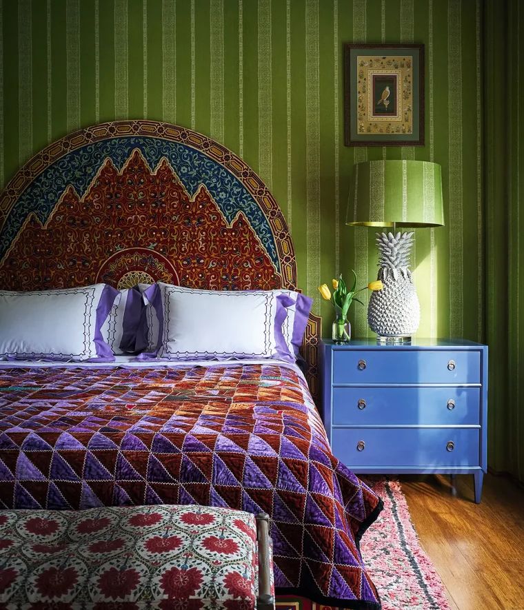 The image shows a neatly made bed with a colorful patterned headboard featuring warm tones and intricate designs A blue nightstand is