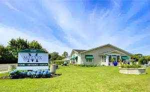 Front view of cozy cottage with green shutters and welcoming porch, set against sunny sky.