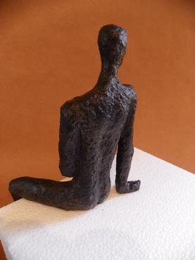 Bronzed metal sculpture of a man sitting with elbow on knee, placed on white surface.