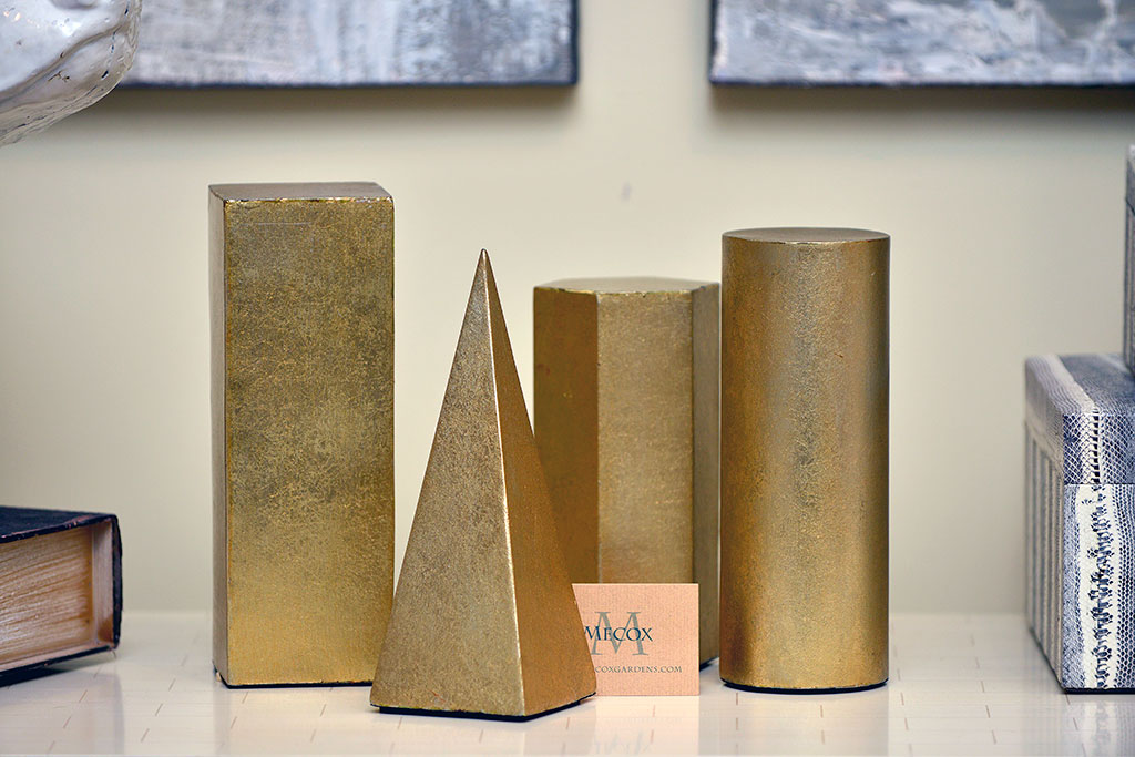 Four brass decorative objects on a shelf: a cylinder, a hexagonal prism, a square pyramid, and a rectangular cuboid.