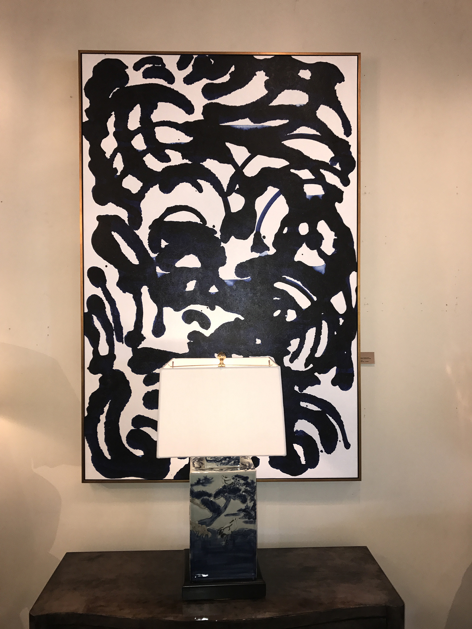 A large abstract painting with a black and white pattern displayed on a wall in a room with a lamp and a