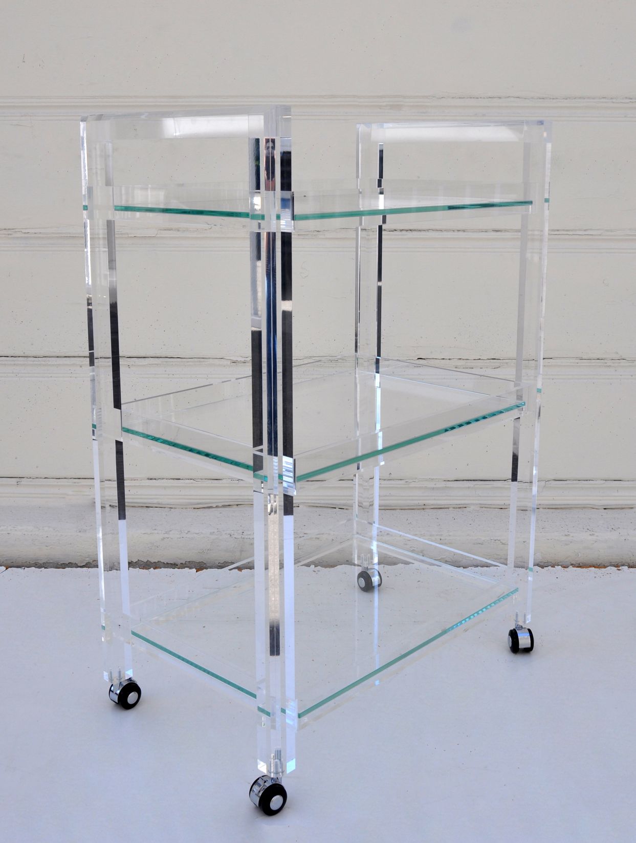 Clear acrylic bar cart with three glass shelves, standing against a white wall.