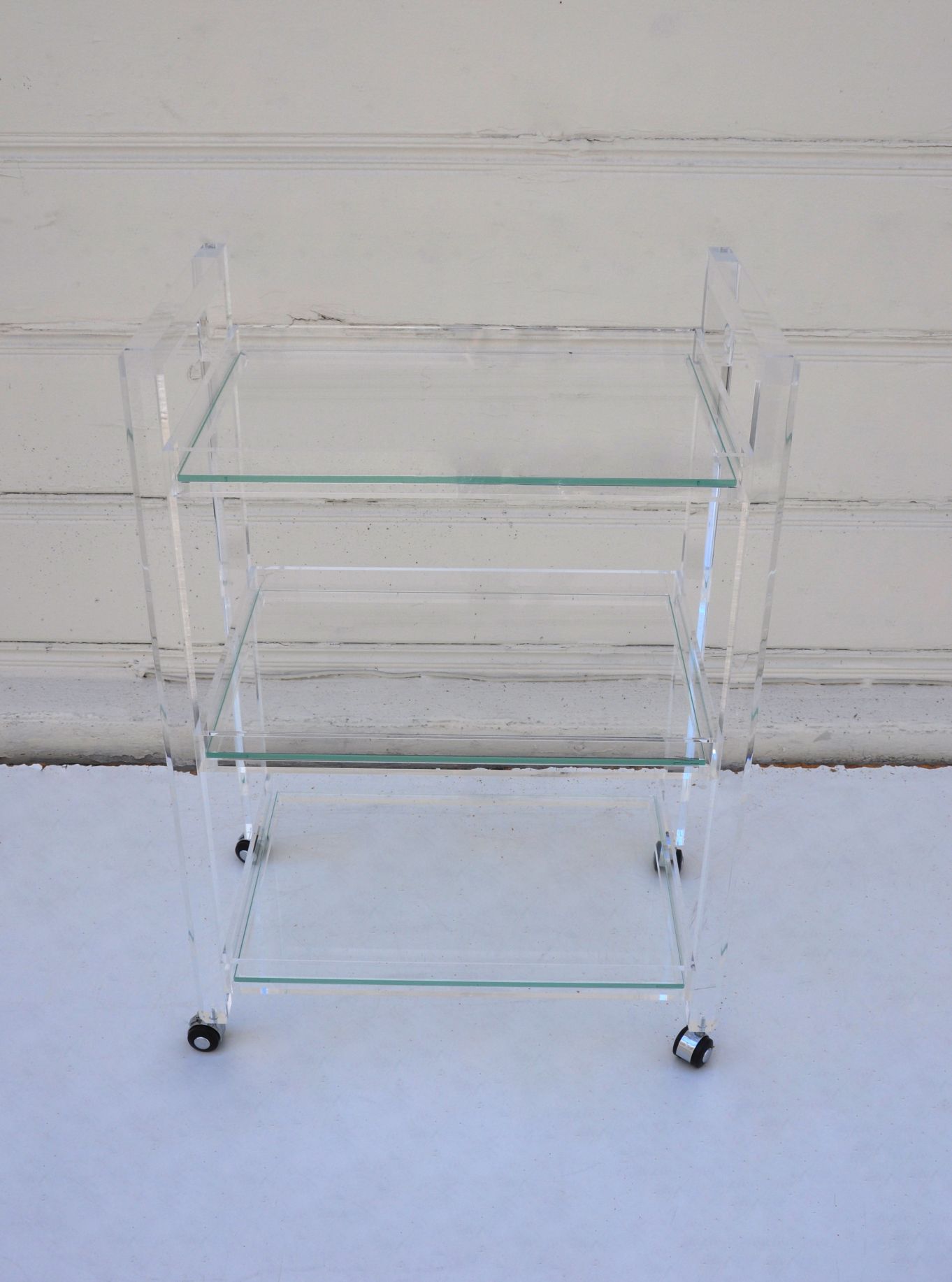 Clear acrylic bar cart with three glass shelves, set against a white wall.