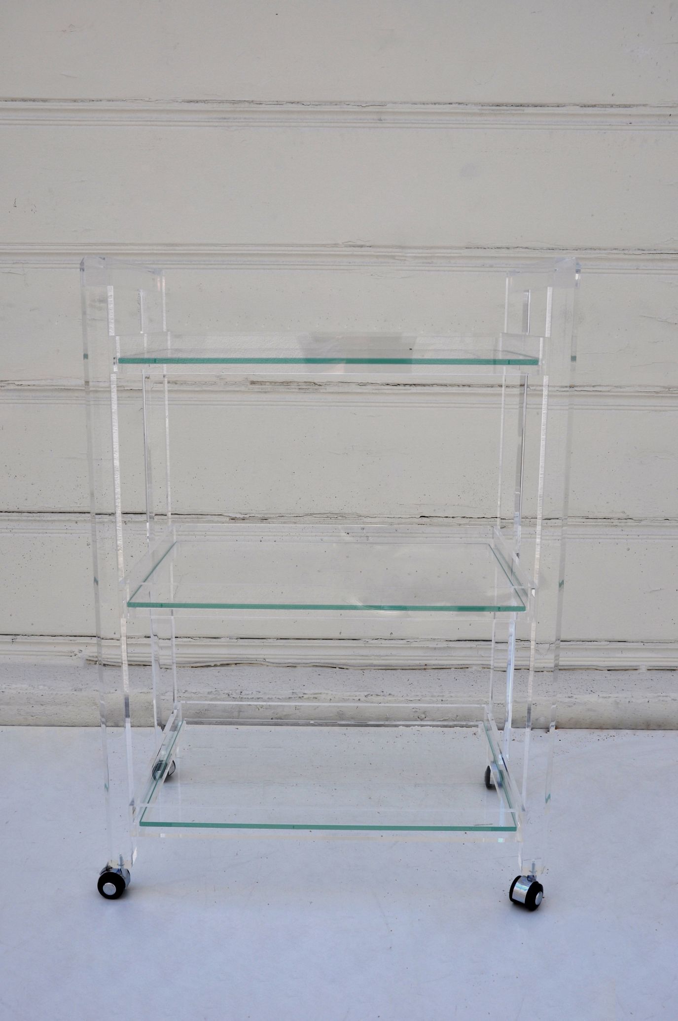 Glass and acrylic bar cart with three shelves, placed against a white wall.