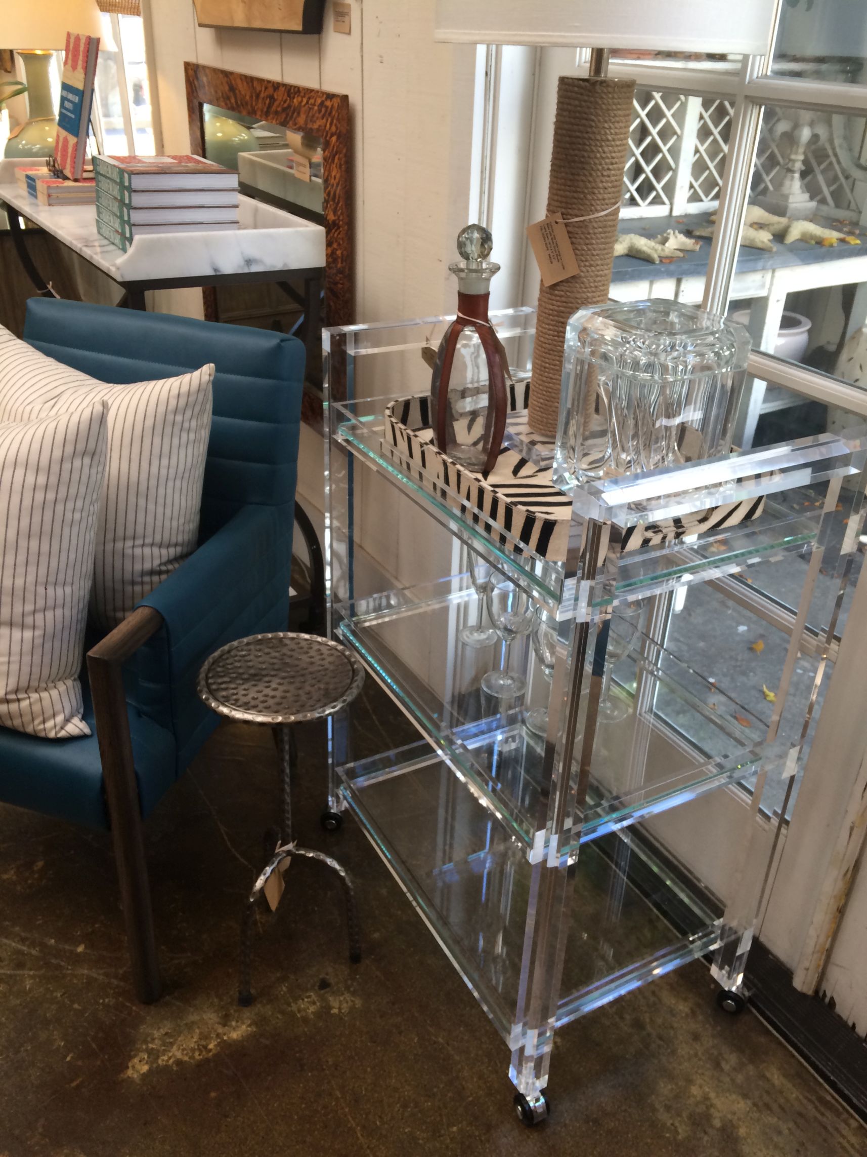 Glass and acrylic bar cart with three shelves, placed in a room with a chair and a table.