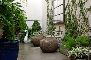 A concrete bench with a wooden appearance, set in an outdoor garden area with potted plants and greenery around it.