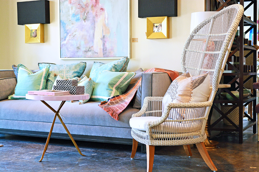A brown wicker chair with a cushion on top, set against a backdrop of a room with a couch and artwork.