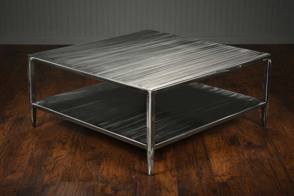Brushed steel coffee table with two shelves and slightly tapered legs, set against a wooden floor in an indoor setting.