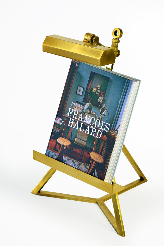 A brass desk lamp with a book on top, placed on a stand against a white background.