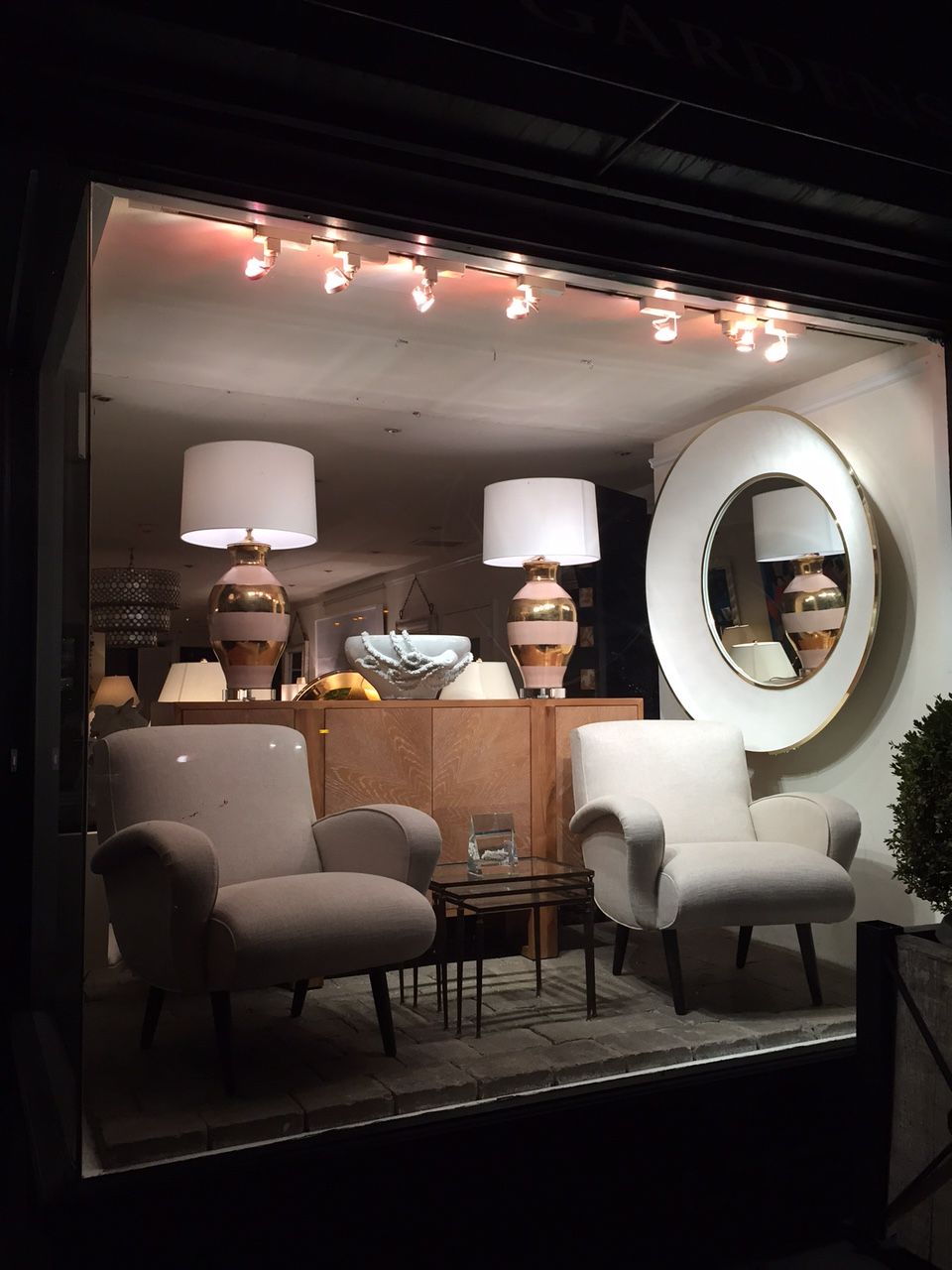 Snow faux shagreen round mirror with a beveled edge and two lamps on either side, displayed in a store window.