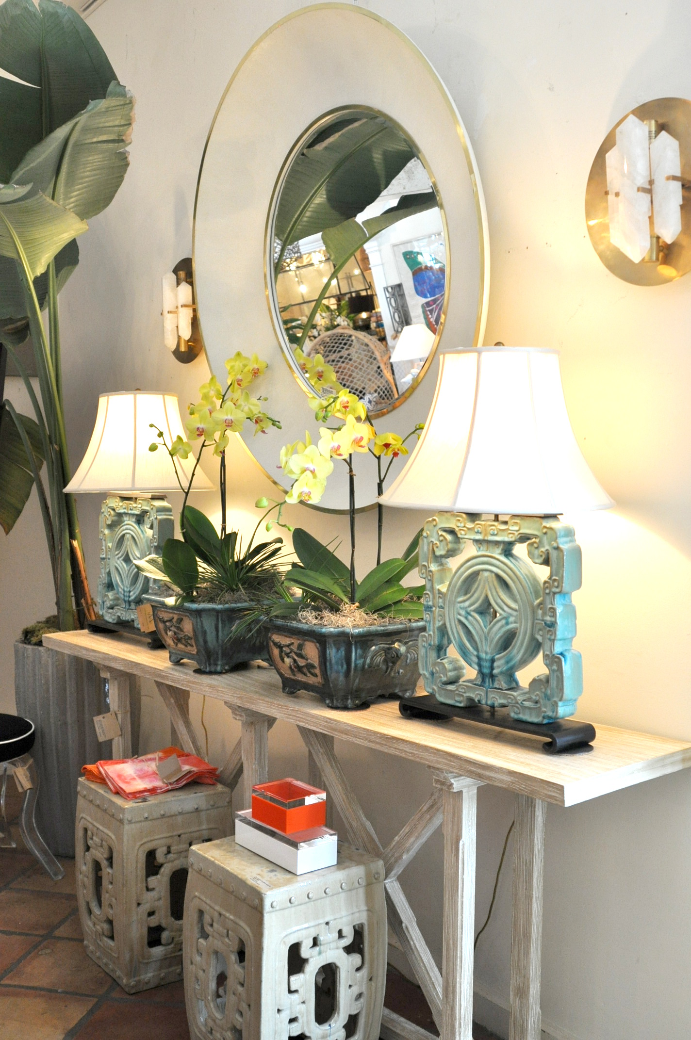 Round mirror with a silver frame on a white wall set against a backdrop of potted plants and a small table with
